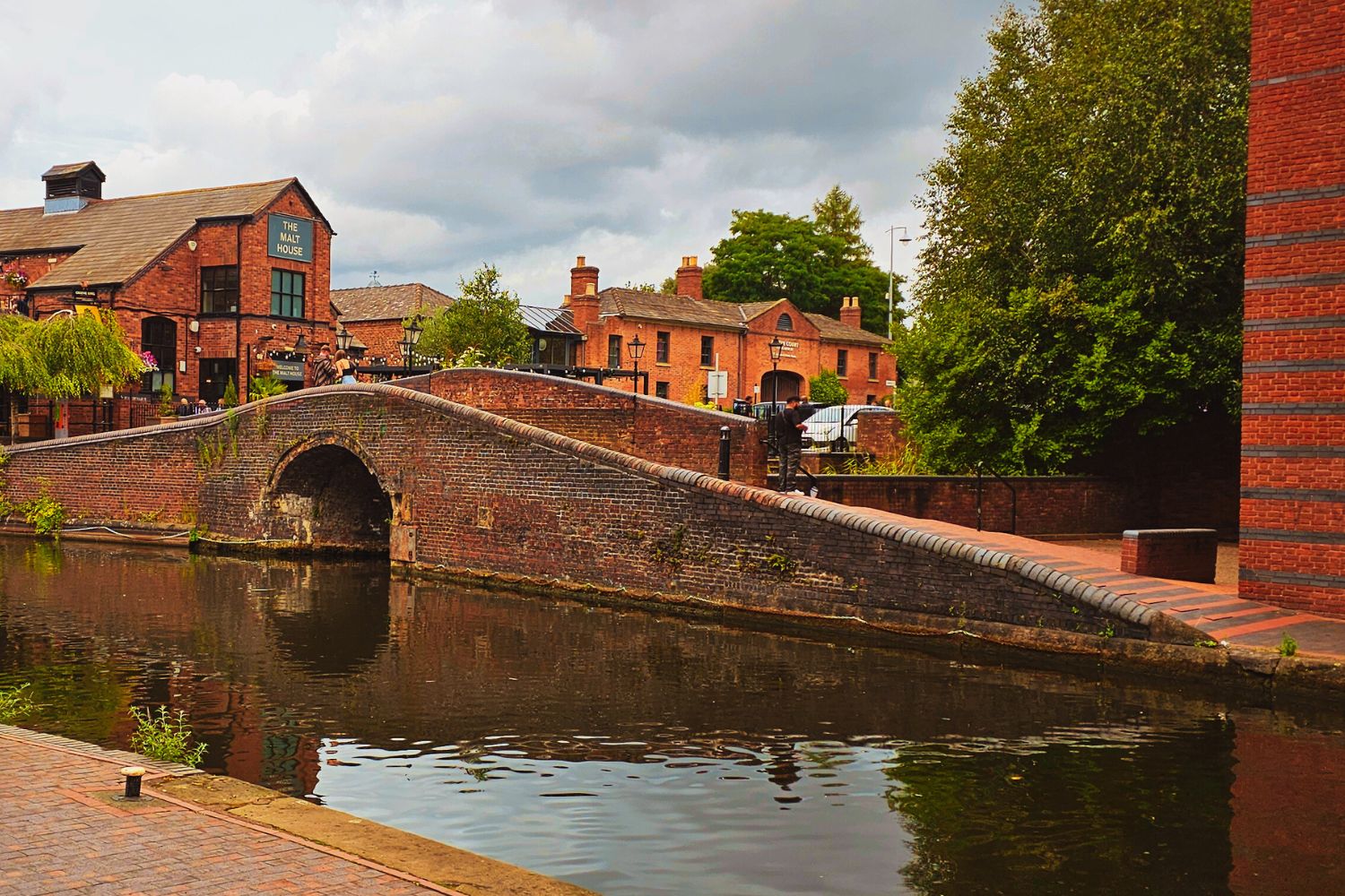 Red-brick bridge with historic buildings in the background and Birmingham Canal in the forefront.