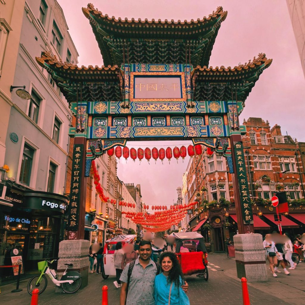 A smiling couple posing in front of the ornate Chinese Gate in London's Chinatown, decorated with rows of traditional red lanterns under a pink sunset sky.