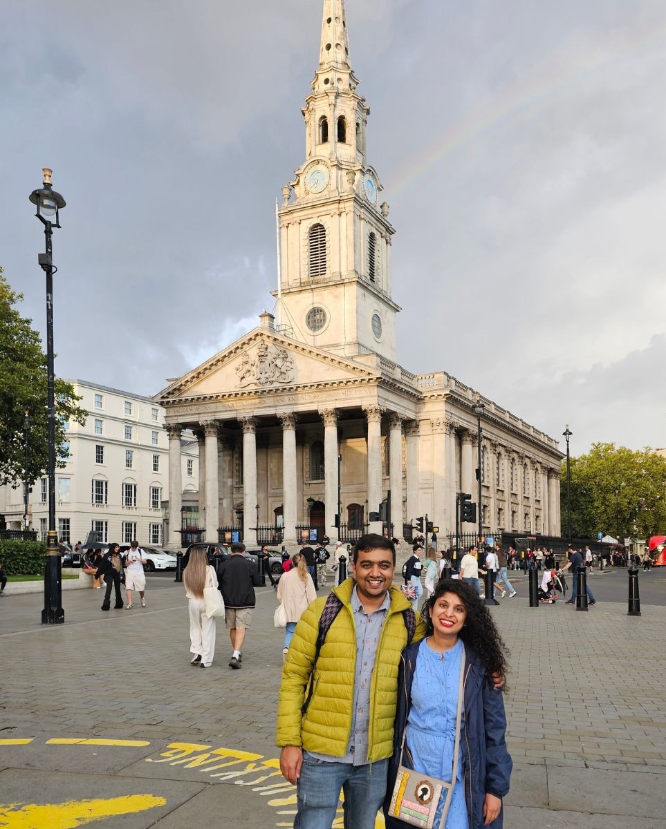 A smiling couple standing in Trafalgar Square, London, with the historic St Martin-in-the-Fields church and a faint rainbow in the sky behind them.