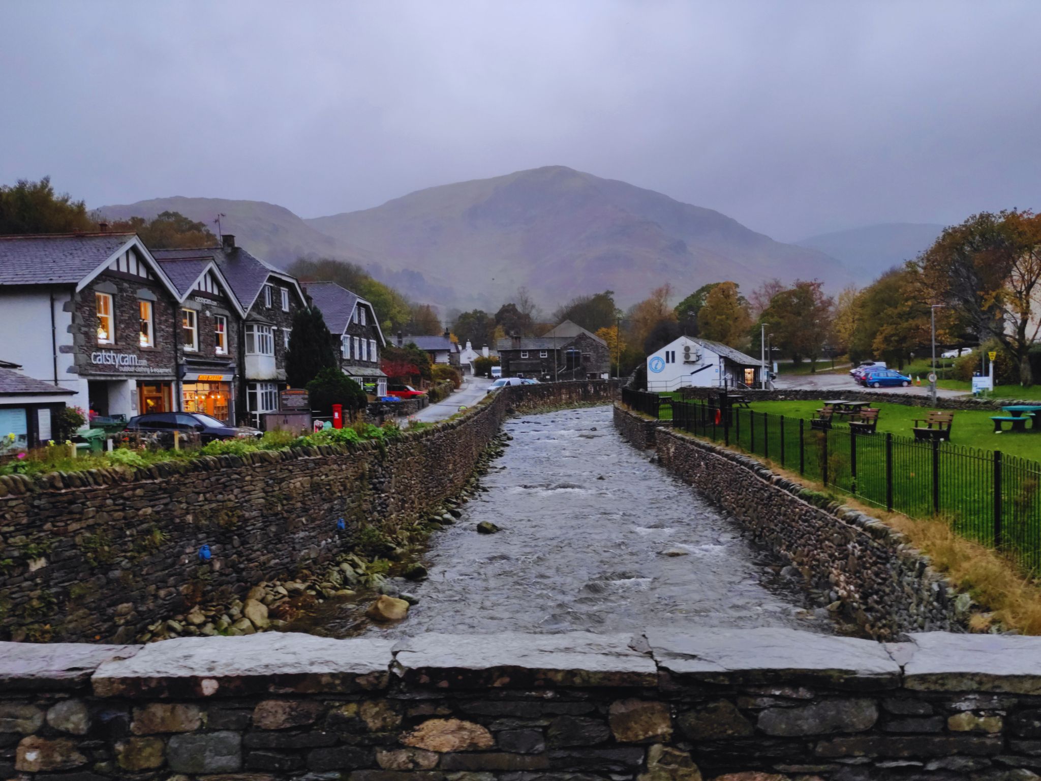 A view from a stone bridge looking down a rocky river (Glenridding Beck) as it flows through the village. To the left, traditional stone-faced buildings with lit shop windows line the bank. To the right, a green park area with benches and a black iron fence. In the background, a massive, misty mountain peak rises into a grey, overcast sky.