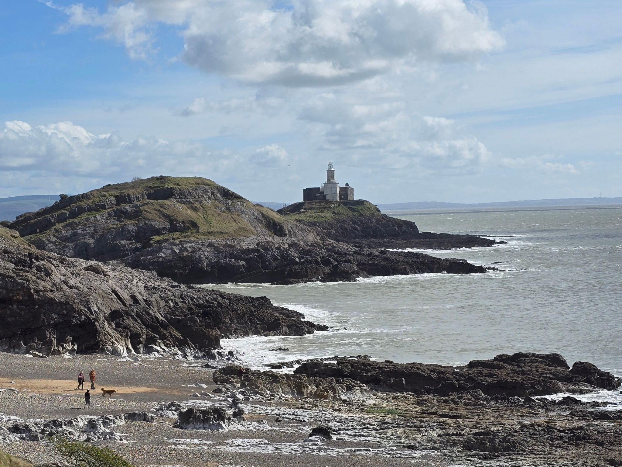 View of the rugged, rocky coastline at Bracelet Bay in South Wales. In the background, the white Mumbles Lighthouse sits prominently on its tidal island. In the foreground, a small sandy and pebbly beach is visible with a few people walking their dog. The sea is grey and choppy, and the sky is blue with dramatic white clouds.