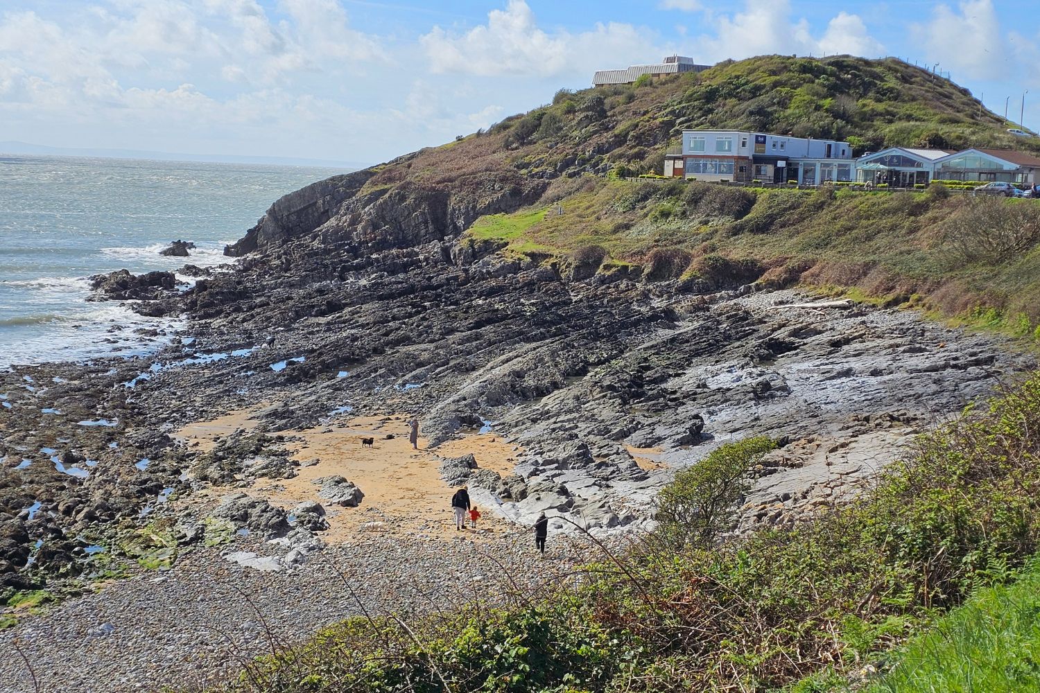 View of the rocky beach and coastline at Limeslade Bay with the Forte’s Ice Cream Parlour and café visible on the hillside above. A few people are walking on the sand and rocks, showing the rugged terrain near the car park area.