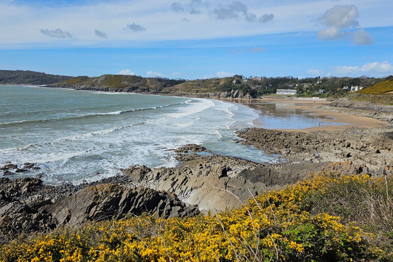 A sweeping view of Caswell Bay from the cliff path, showing the mix of jagged rocks, sandy beach, and distant white coastal buildings.