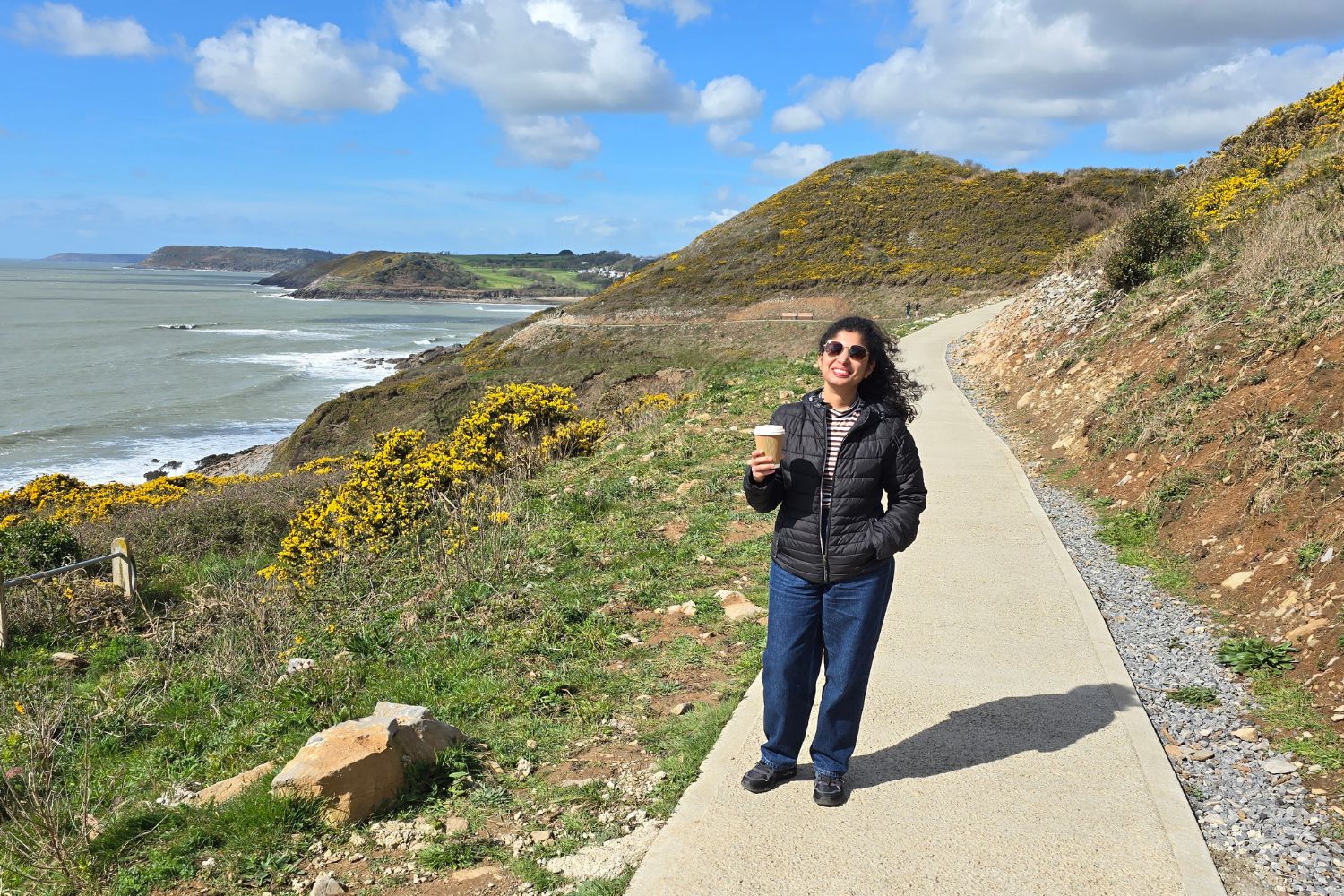 A smiling hiker in a black quilted jacket and sunglasses stands on a smooth, paved section of the Wales Coast Path. She is holding a takeaway coffee cup, with bright yellow gorse bushes, rolling green hills, and the blue ocean of the Gower Peninsula stretching out behind her under a sunny sky.