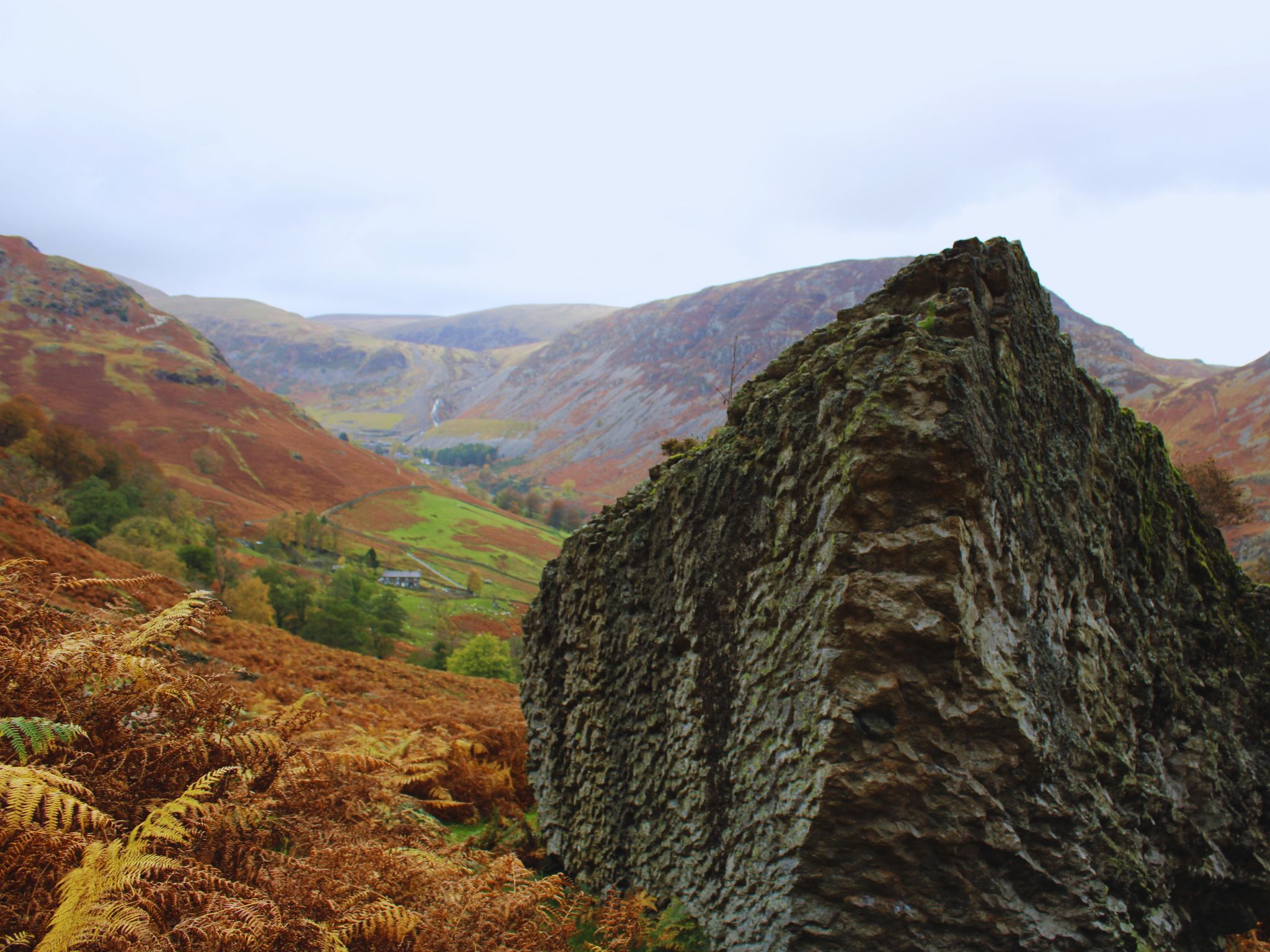A large, jagged grey rock with deep vertical ridges sits in the foreground on a hillside. Beyond the rock, the view opens up to a deep valley with green fields, scattered stone houses, and steep, copper-coloured mountainsides under a cloudy sky.