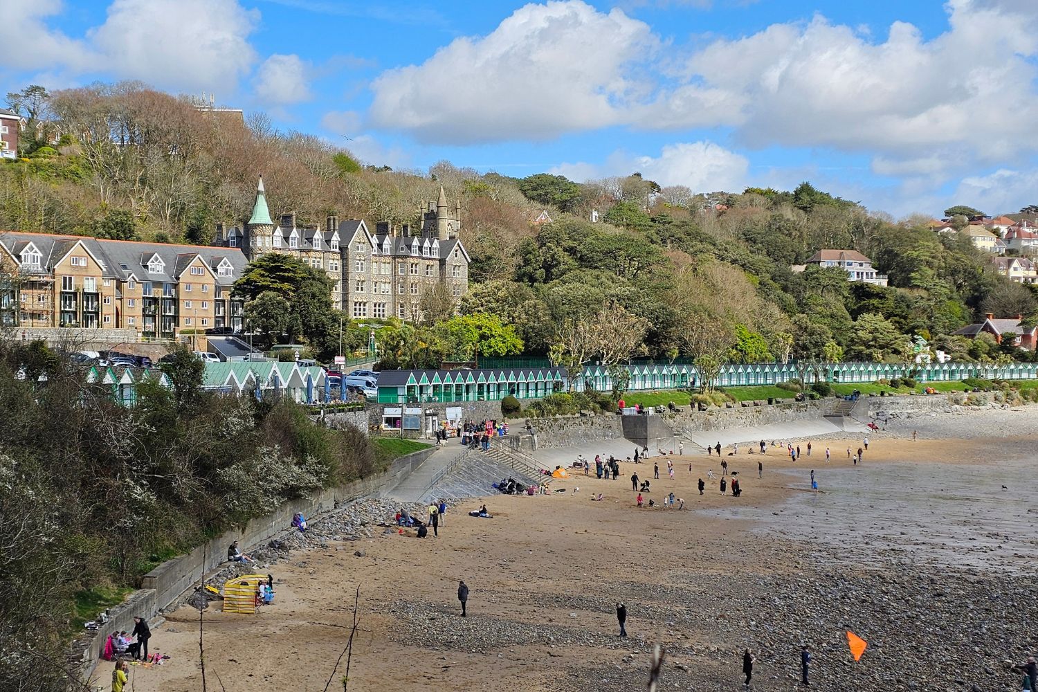 A panoramic view of Langland Bay in Gower, Wales, featuring the iconic long row of green and white beach huts along the promenade. In the background, the grand, turreted Langland Manor stands on the hillside. The beach shows a mix of golden sand and pebbles with many visitors enjoying the shoreline under a bright, cloudy sky.