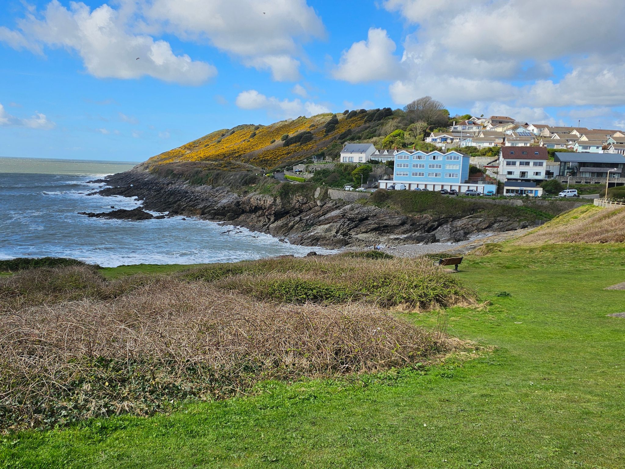 A wide view of Limeslade Bay featuring a prominent light-blue building on the cliffside. The surrounding hills are covered in vibrant yellow gorse, and the rocky coastline meets the choppy sea under a bright, cloud-filled sky.