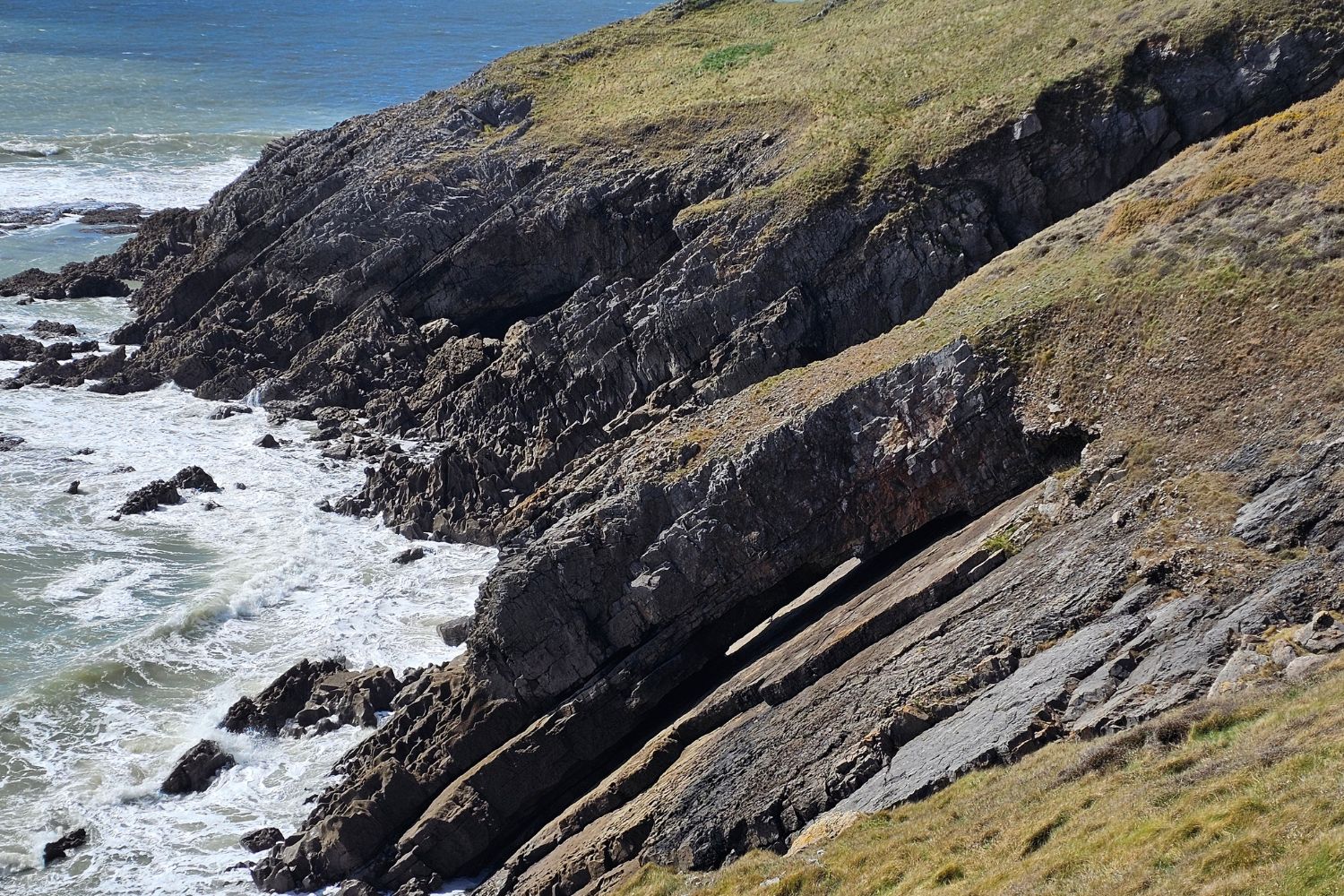 A close-up view of the rugged limestone cliffs near Langland Bay, showing deep, dramatic gaps and horizontal erosion lines in the soft stone. The churning white sea waves crash against the base of the dark, weathered rock formations.
