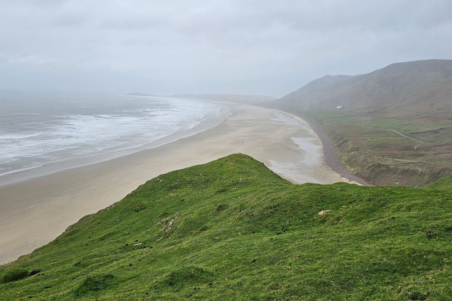 A moody, high-angle view of the vast Rhossili Bay during a storm. The wide sandy beach and rolling green hills are shrouded in grey mist and low clouds, with white waves rolling onto the shore from a dark sea.