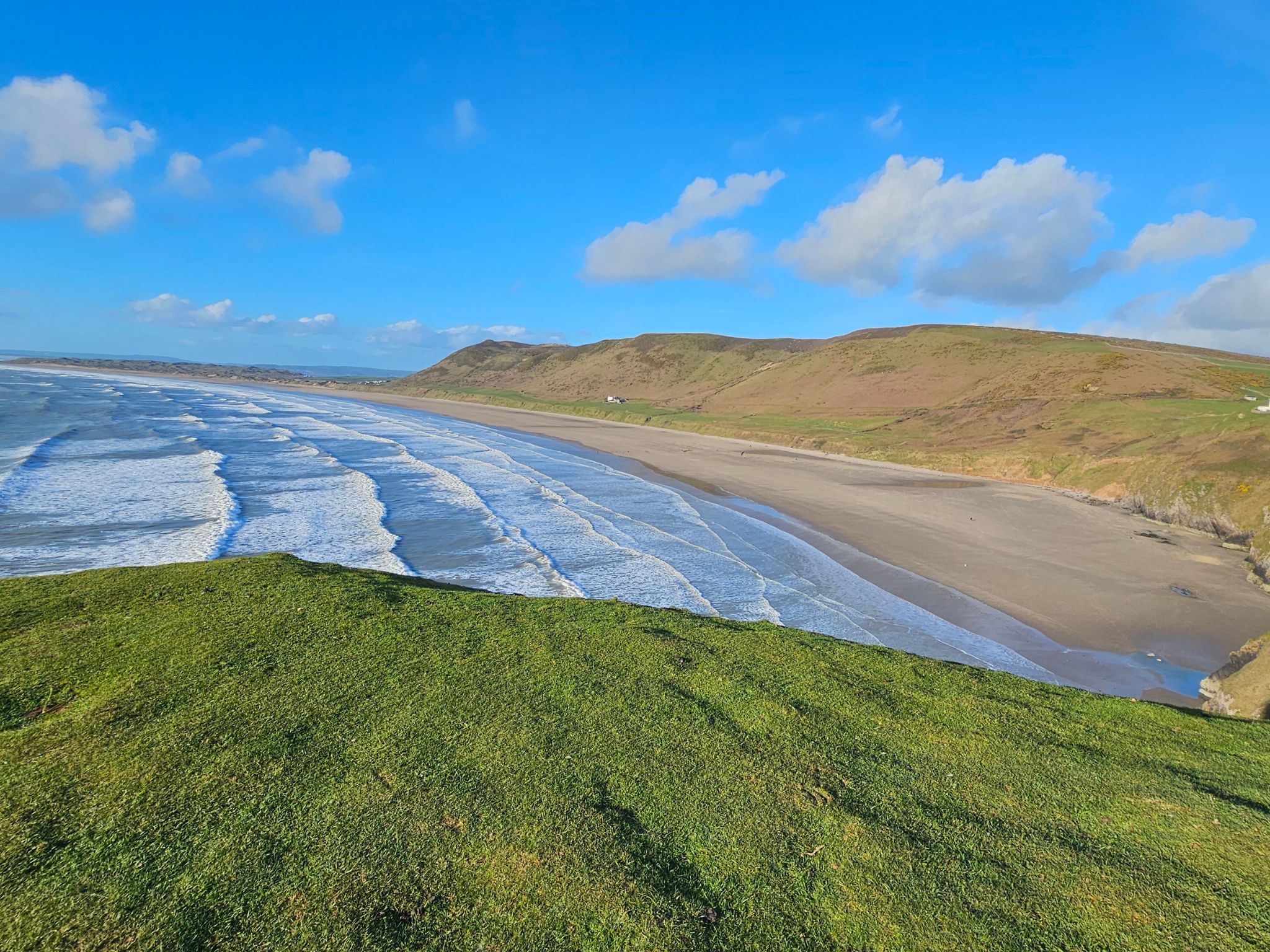 High-angle panoramic view of the vast, crescent-shaped Rhossili Bay beach with white waves rolling onto the sand and green clifftops in the foreground under a bright blue sky.