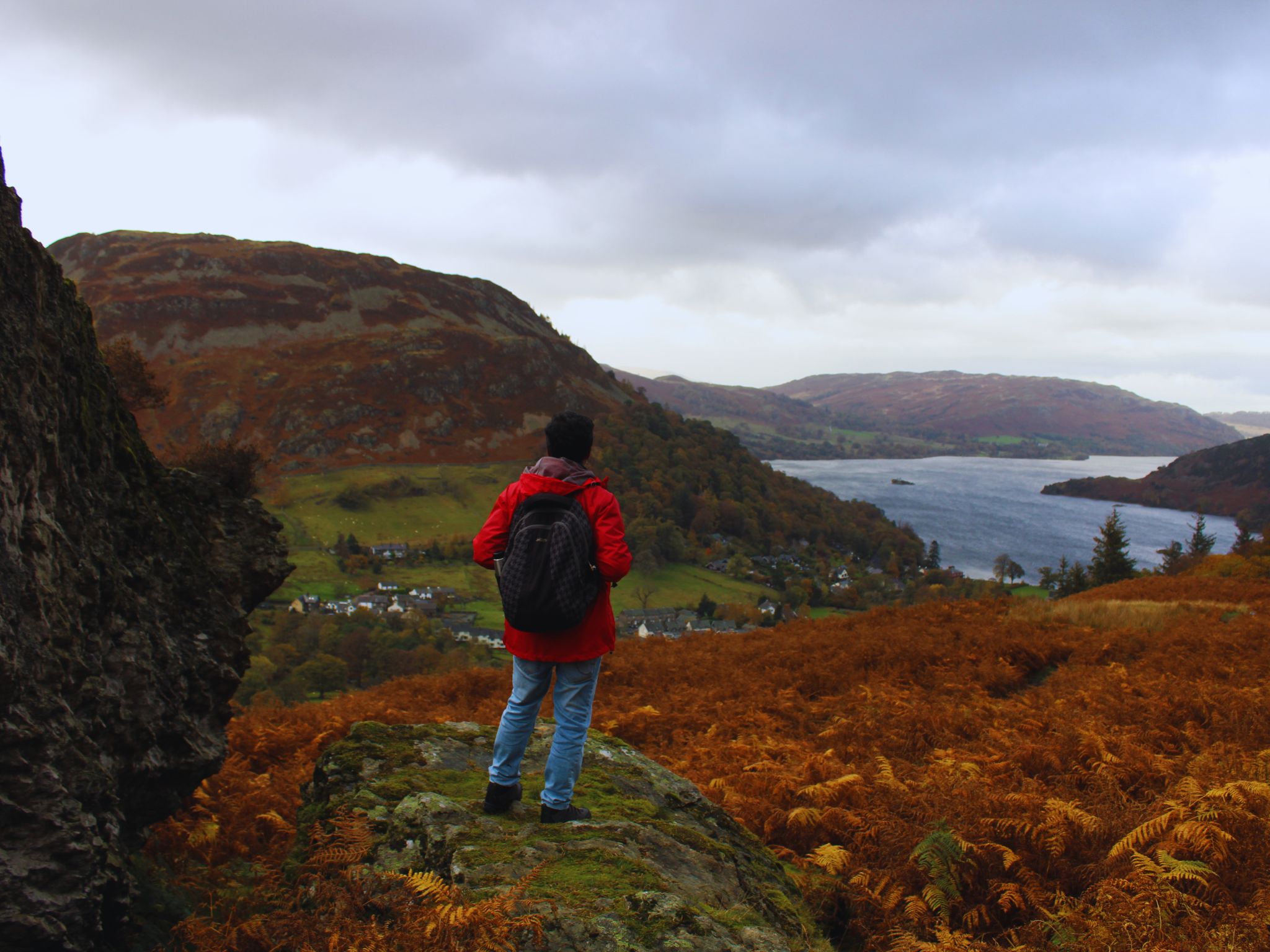 A hiker in a bright red jacket and backpack stands on a rocky, mossy outcrop, looking out over Ullswater. To the left is a large, dark rock face. Below them, a sea of orange autumn bracken leads down to the village of Glenridding and the long, blue lake under a dramatic, cloudy sky.