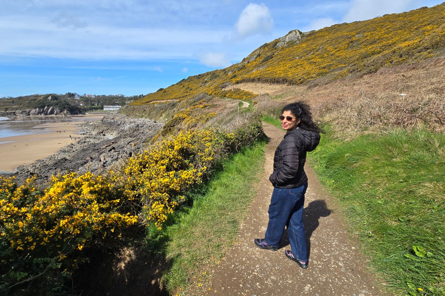 A traveller stands on a narrow, stony section of the Gower Coastal Path, looking back over their shoulder. The ground underfoot is uneven and covered in small grey rocks, illustrating the change in terrain. The hillside is covered in bright yellow gorse, with the sandy shoreline of Caswell Bay visible in the distance.