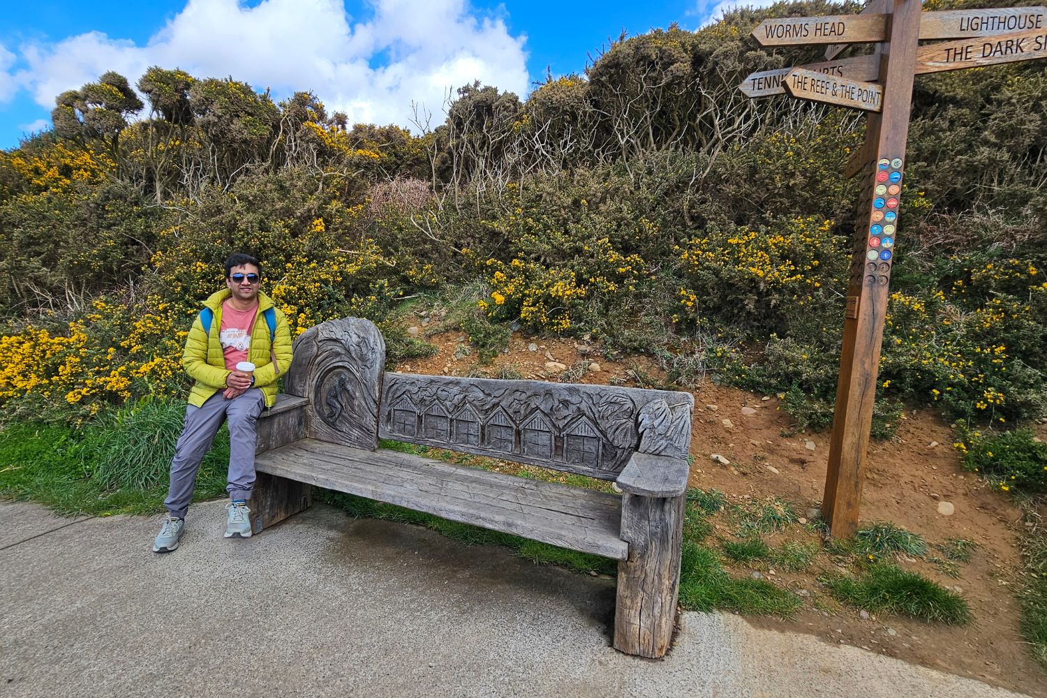 A traveller sitting on the unique carved wooden "Surfer Bench" on the Gower Coastal Path. The bench features detailed carvings of a surfer and beach houses, set against a backdrop of vibrant yellow gorse and a wooden signpost pointing toward Worms Head and the Lighthouse.