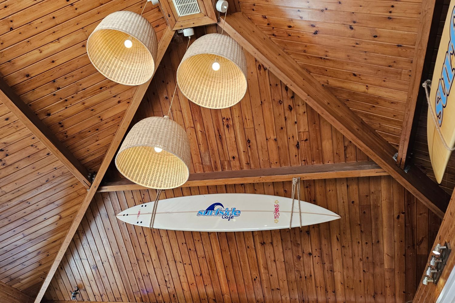 The interior of Surfside Café at Caswell Bay, showing a high vaulted wooden ceiling with warm lighting. Three large woven wicker pendant lamps hang from the beams, and a white surfboard with the "Surfside Café" logo is suspended as décor.
