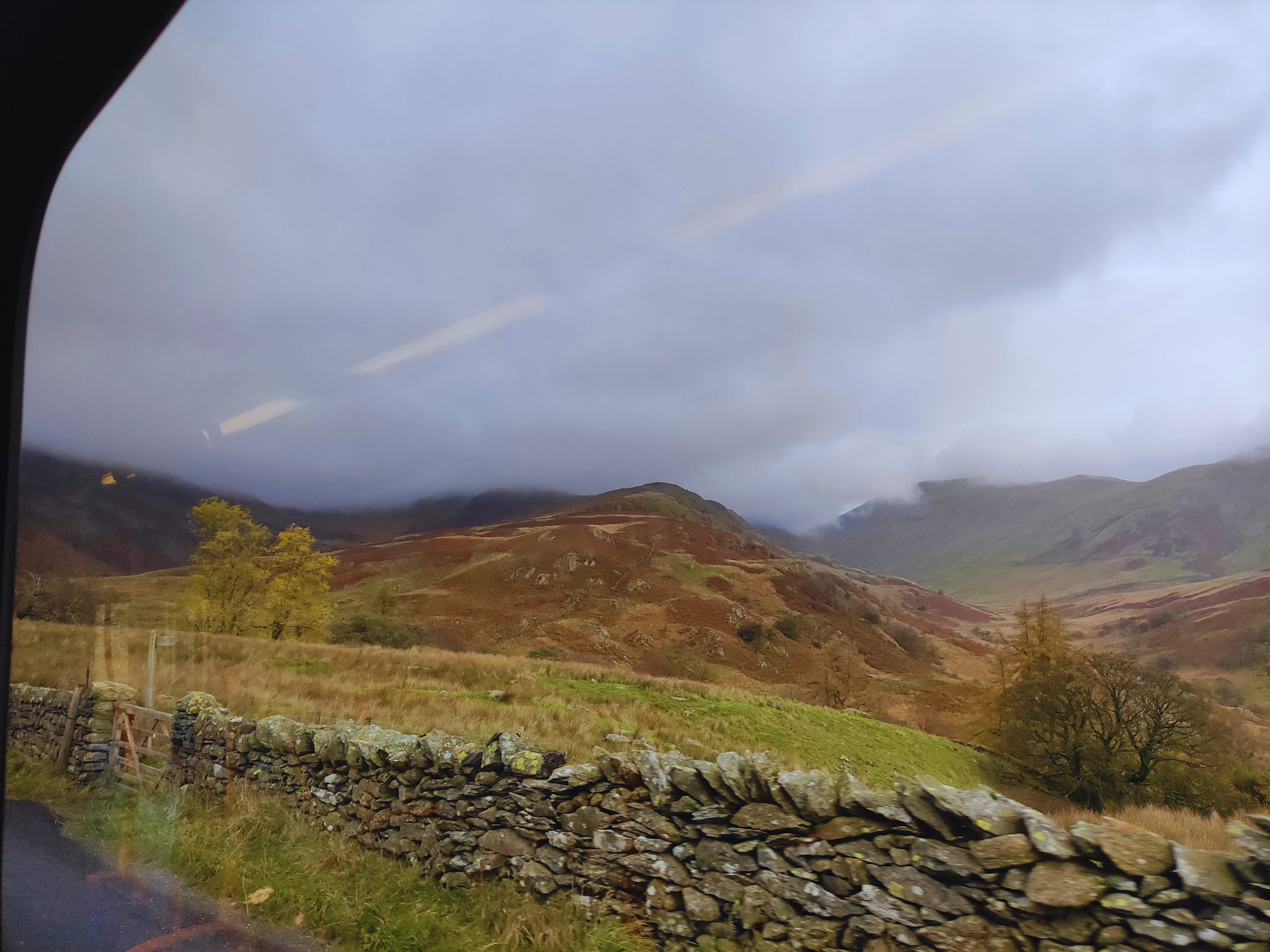 A view from a bus window looking out over the rolling, misty fells of the Lake District. In the foreground, a traditional dry-stone wall and a wooden gate blur past. In the distance, brown and green hills rise into a heavy, low-hanging grey sky, with clouds touching the mountain peaks.