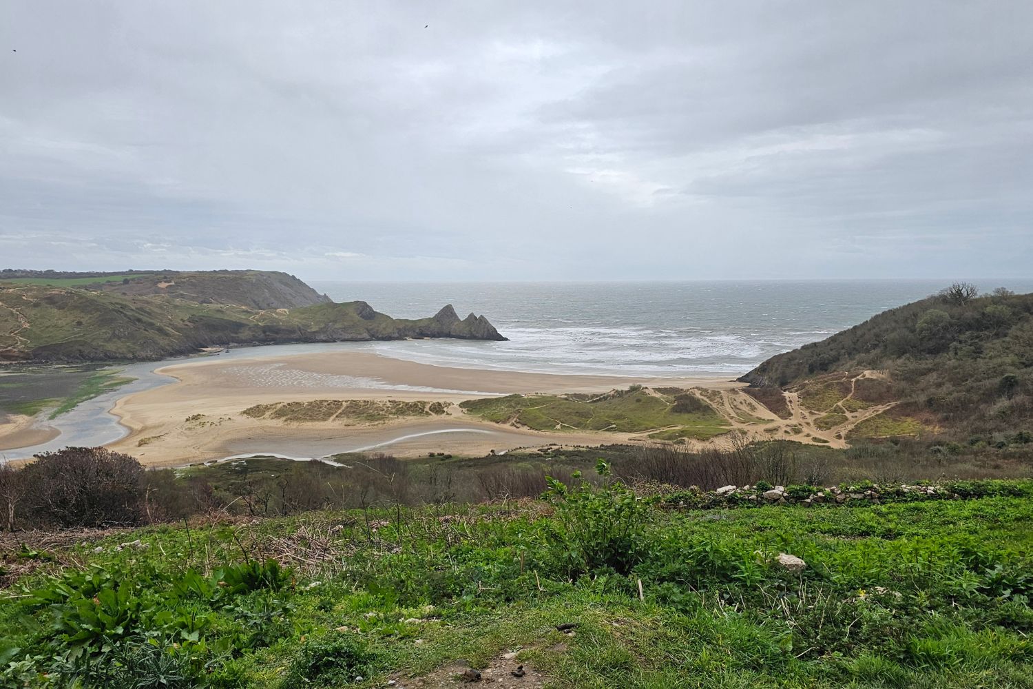 A wide, high-angle view of Three Cliffs Bay under an overcast sky. The iconic trio of jagged limestone peaks sits at the edge of the sandy shoreline where the river meets the sea, surrounded by green dunes and rugged cliffs.