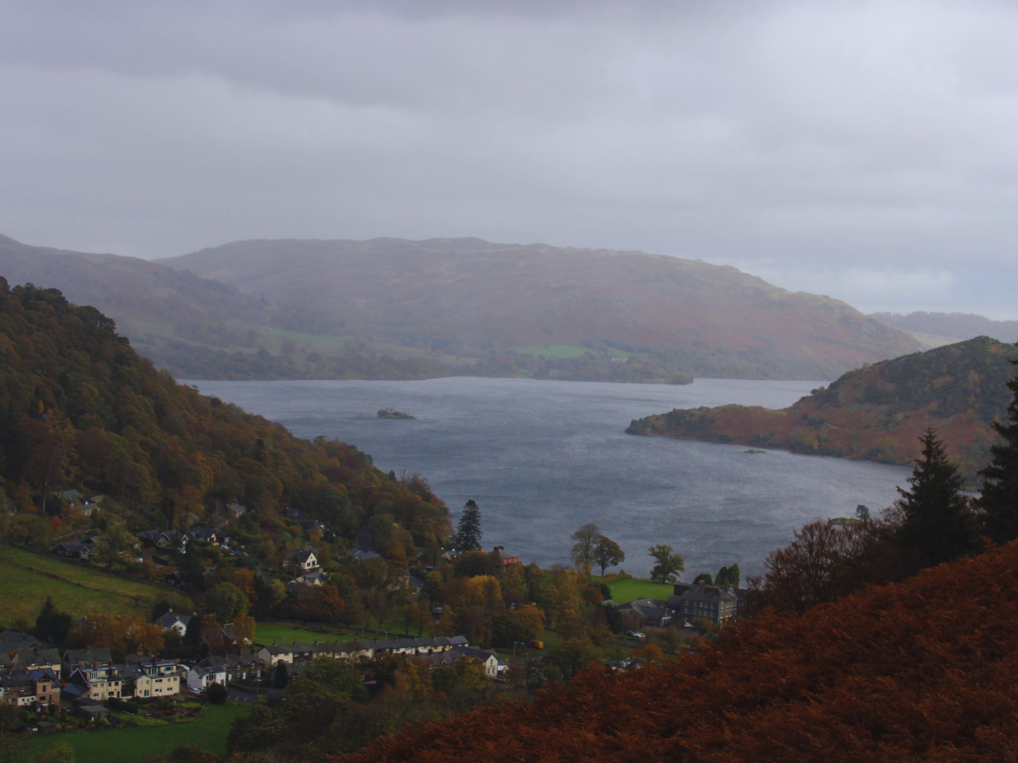 A wide, high-angle view of Ullswater lake from a mountain trail. In the valley below, the cluster of white and grey buildings of Glenridding village sits among green fields and autumn trees. The deep blue lake is framed by steep, rolling fells under a heavy, grey overcast sky.