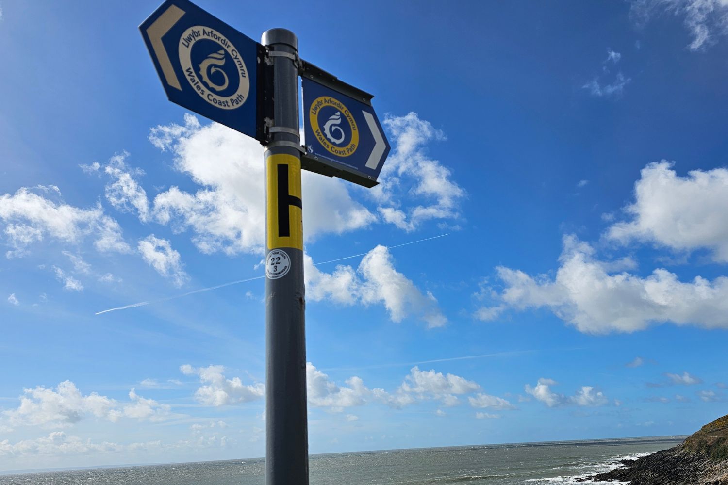 A close-up of a Wales Coast Path signpost against a bright blue sky. The blue signs feature the yellow and white nautilus shell emblem (the "snail" symbol) and the text "Llwybr Arfordir Cymru - Wales Coast Path."
