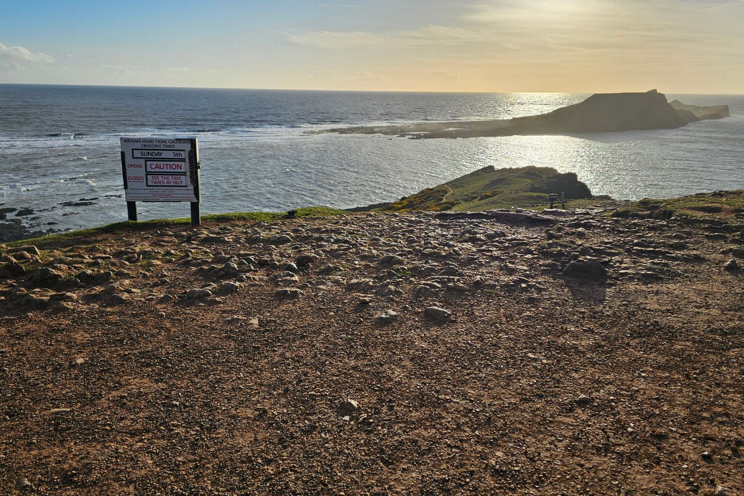A wide-angle shot of a warning sign on a rocky clifftop at Rhossili, overlooking the sea and the Worm's Head tidal island. The sign displays the date "Sunday 5th" and a large red "CAUTION" warning, stating that the causeway is closed and advising visitors to check tide times at the hut. The sun sets low over the water, casting a golden glow across the landscape.