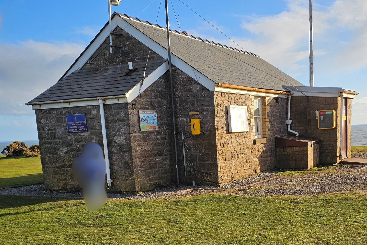 A small, sturdy stone building known as the Worms Head Coastwatch Station, featuring a grey slate roof and several information plaques on its exterior. The station sits on a grassy clifftop under a clear blue sky, overlooking the sea on the Gower Peninsula.