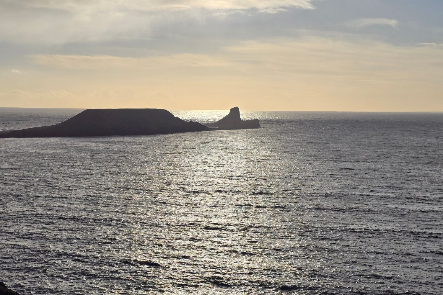 A wide silhouette of Worm's Head, a jagged tidal island, stretching into the shimmering sea at sunset. The sunlight reflects off the calm water, creating a bright path of light toward the dark, rocky headland under a soft, hazy sky.