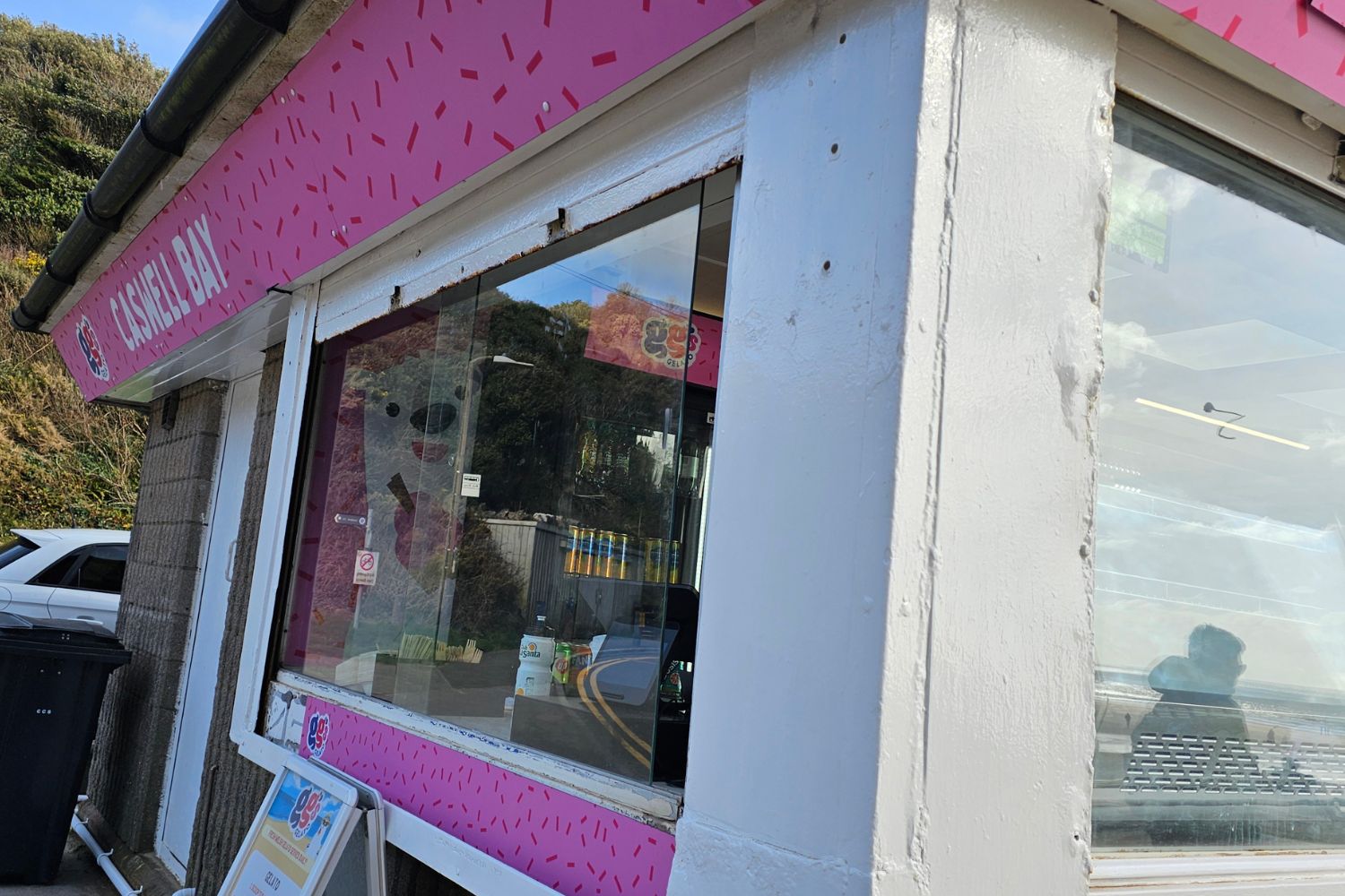 Close-up of the bright pink "GG's Gelato" ice cream stand at Caswell Bay. The shop has a white exterior with pink sprinkle-patterned signage, reflecting the coastal road and scenery in its glass window.